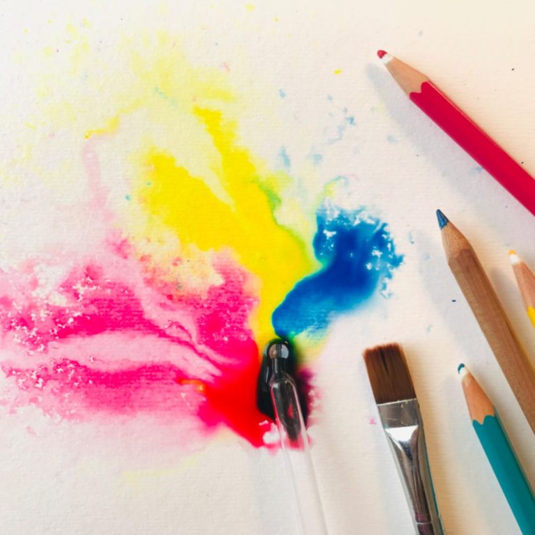 Rote, gelbe und blaue Farbkleckse auf weißem Papier, daneben liegen Stifte und Pinsel. 